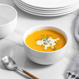 Bowl of pumpkin cream soup with a spoon and white plates on a white table