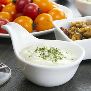 White bowl of creamy dip with herbs on a table with tomatoes and other food.