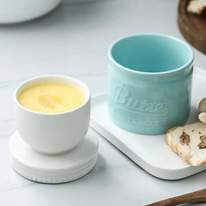 Two ceramic containers, one white with a yellow substance and the other light blue with 'Butter' text, on a light surface with bread slices.