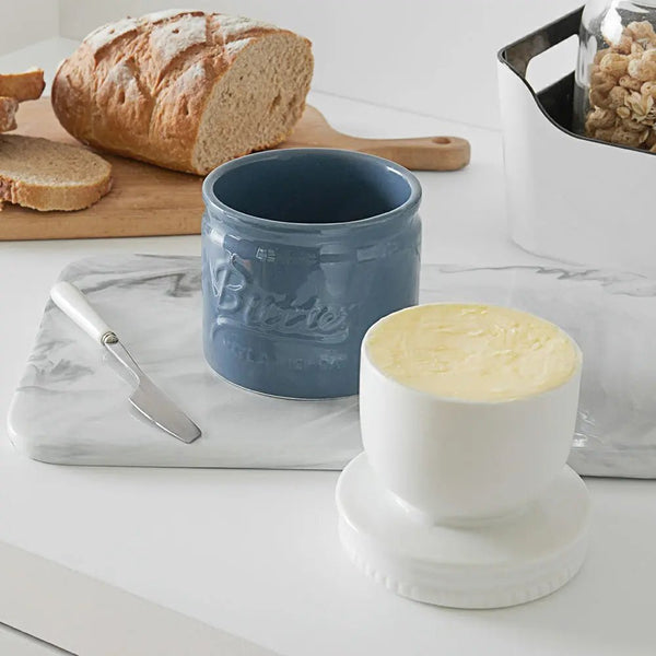 Blue ceramic crock and white ceramic crock with lid on a marble surface with bread and a knife.