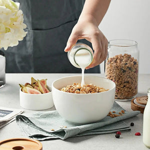 Person pouring milk into a bowl of granola on a table with a jar of milk and a bowl of fruit in the background.