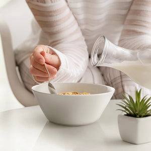 Person pouring milk into a bowl of cereal on a white surface with a plant in the background.