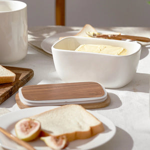 White ceramic butter dish with wooden lid on a table with bread and figs.