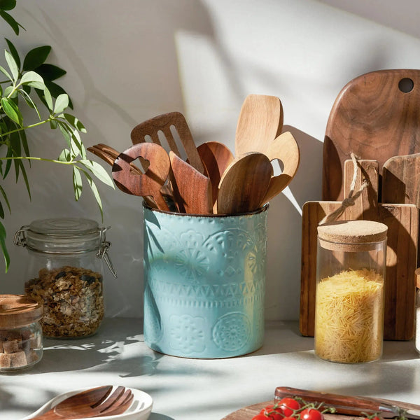 Kitchen utensils in a blue container with wooden cutting boards and jars on a light surface.