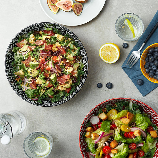 Two bowls of salad with various ingredients on a table with lemon, figs, and blueberries.