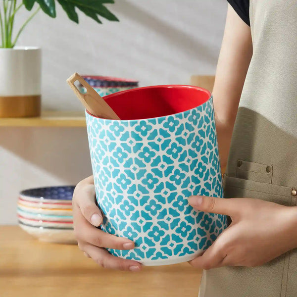 Person holding a blue patterned ceramic utensil holder with a red interior, kitchen setting