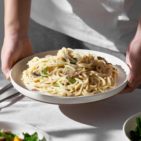 Person holding a plate of pasta with vegetables on a light surface