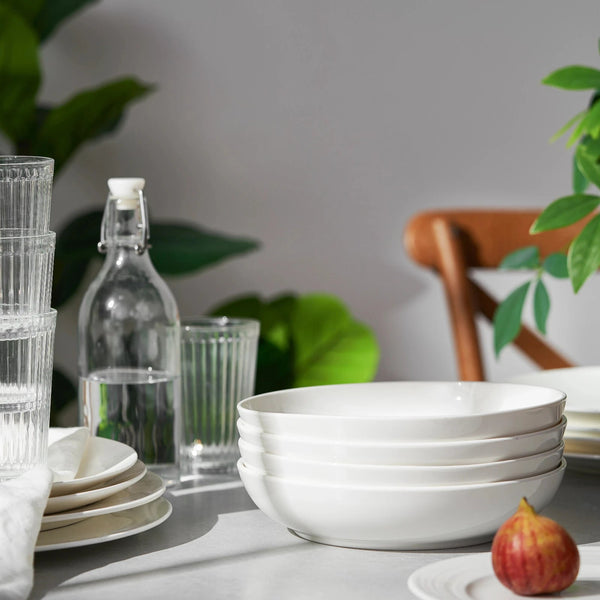 Stack of white bowls on a table with glasses, a bottle, and a chair in the background.