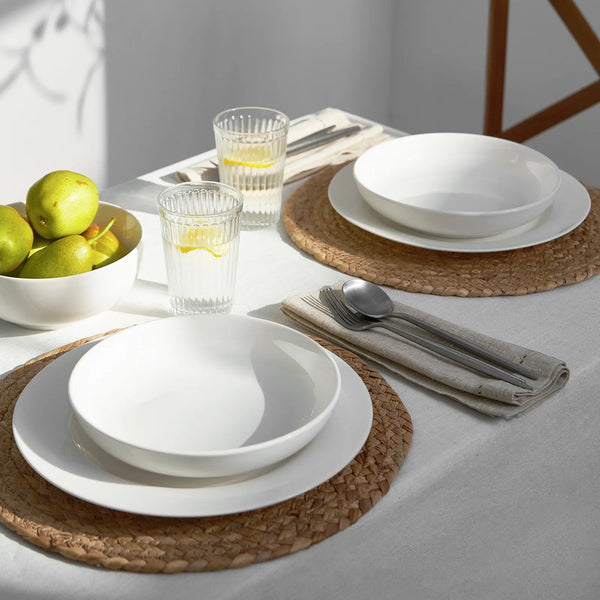 Dining table setting with white plates, glasses, and cutlery on woven placemats.