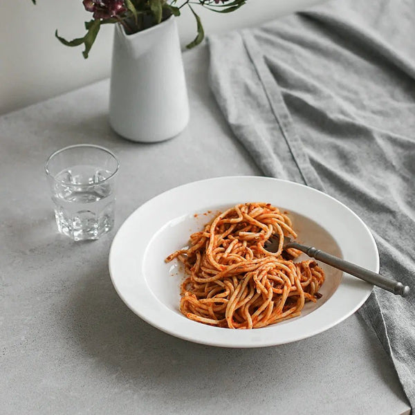 White plate with spaghetti, a glass of water, and a vase with flowers on a gray surface.