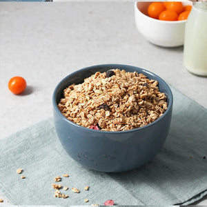 Blue bowl of granola on a light gray surface with a white bowl of oranges and a glass of milk in the background.