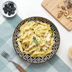 Pasta dish on a patterned plate with a side of blueberries and nuts on a checkered tablecloth.