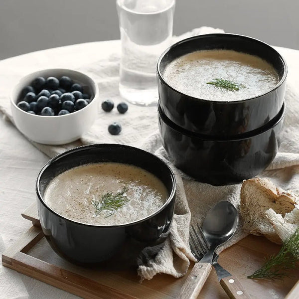 Two black bowls of soup with a side of bread and blueberries on a table.
