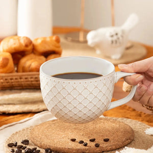 White mug with gold patterns filled with coffee on a cork coaster, with pastries in the background.