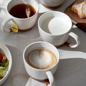 large ceramic coffee cups containing coffee, tea, and a latte art design on a light gray surface.