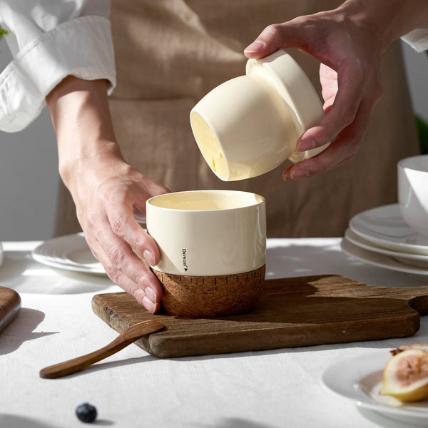 Person pouring a white ceramic cup into another on a wooden cutting board with a neutral background.