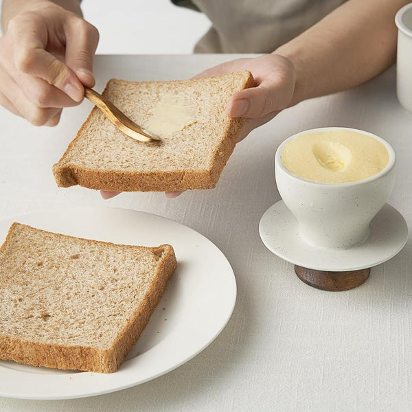 Person spreading butter on a slice of bread with a knife, next to a bowl of butter and another slice of bread on a white plate.