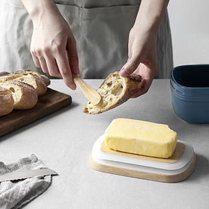 Person spreading butter on bread with a wooden knife, surrounded by bread and a blue bowl on a light gray surface.