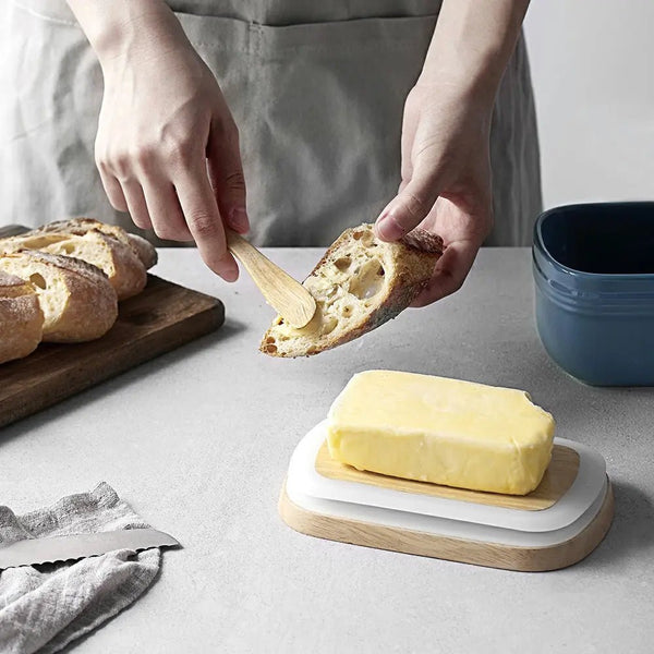 Person spreading butter on bread with a wooden knife, surrounded by bread and a blue bowl on a light gray surface.