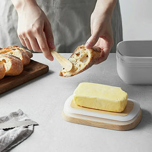 Butter dish with a stick of butter on a wooden board, with bread being cut into and a person holding a piece.
