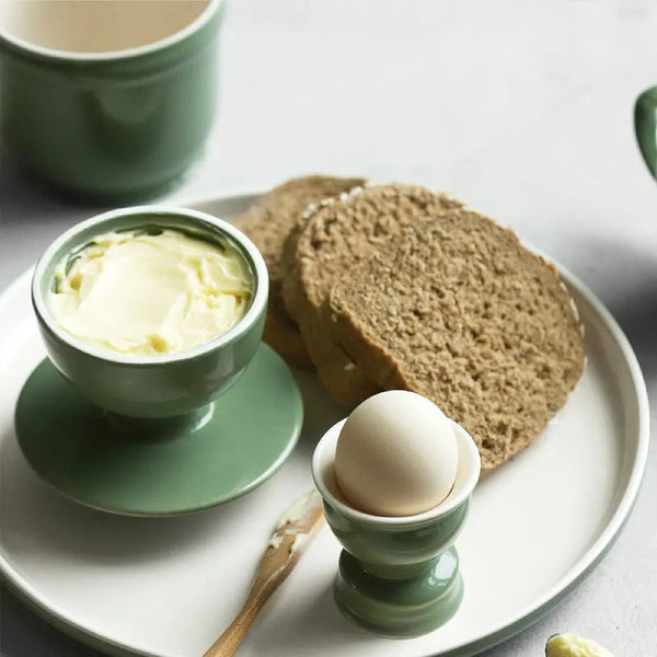 Green ceramic bowl with butter, egg in a cup, and slices of bread on a white plate.