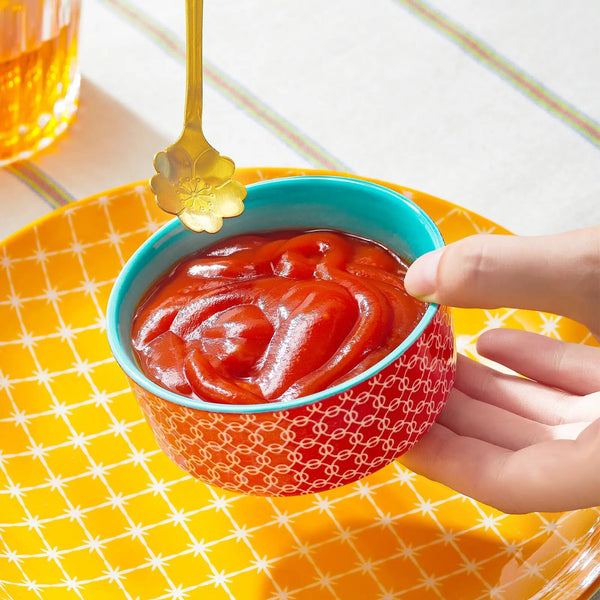 Hand dipping a golden spoon into a bowl of ketchup on a yellow checkered tablecloth.