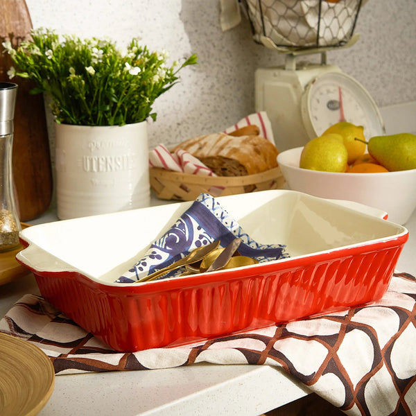 Red ceramic dish on a kitchen counter with a plant, bread, and fruit in the background.