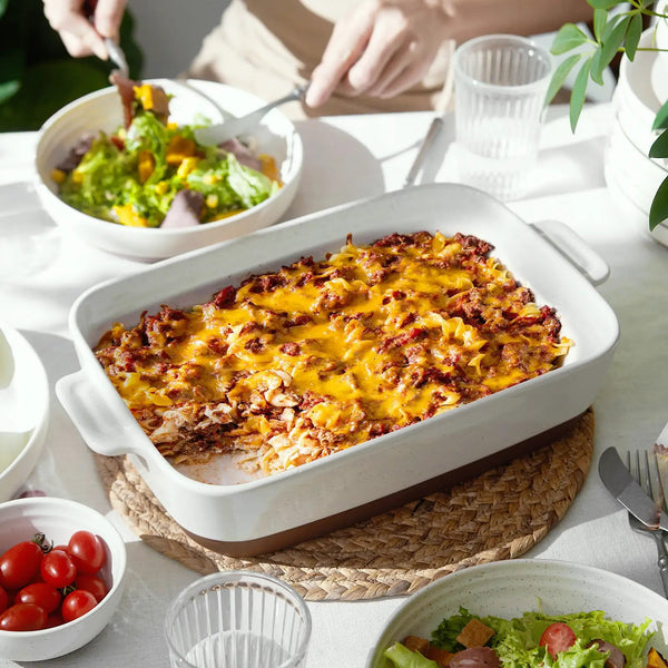 Baked dish in a white casserole dish on a table with salad and tomatoes.