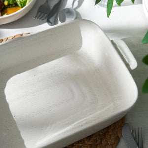 White ceramic dish on a table with a salad and cutlery in the background