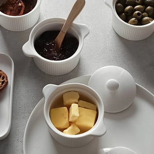 White ceramic ramekins containing butter and a dark spread, with a wooden spoon, on a light gray surface.