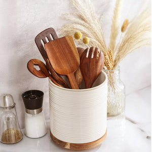 Wooden kitchen utensils in a white container on a light surface with decorative elements.