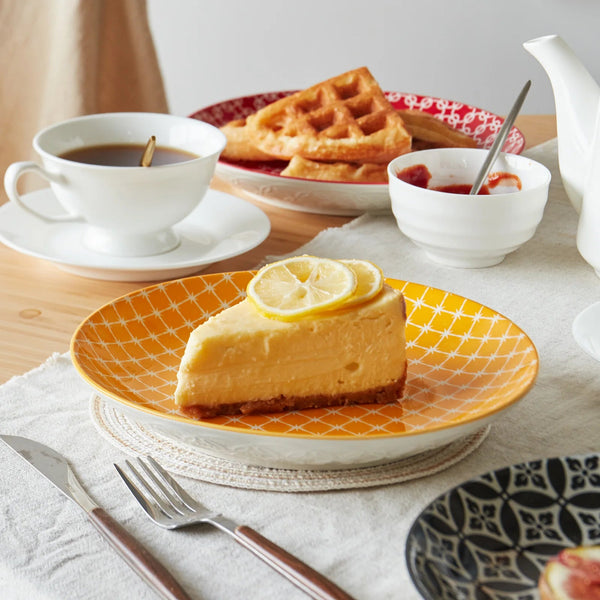 Slice of lemon cheesecake on an orange plate with a cup of coffee and waffles in the background.