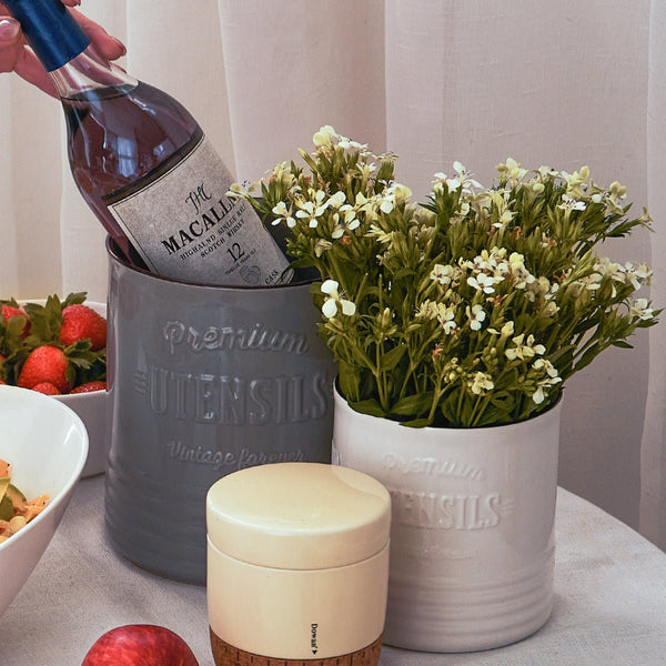Gray container with 'Butlers' branding, bottle of wine, white vase with flowers, and fruit on a table.