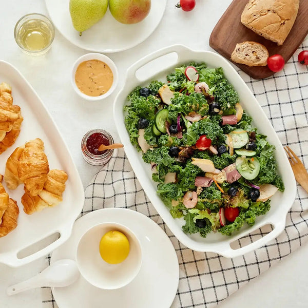 Salad with greens, tomatoes, and black olives on a white dish, surrounded by pastries and fruit.
